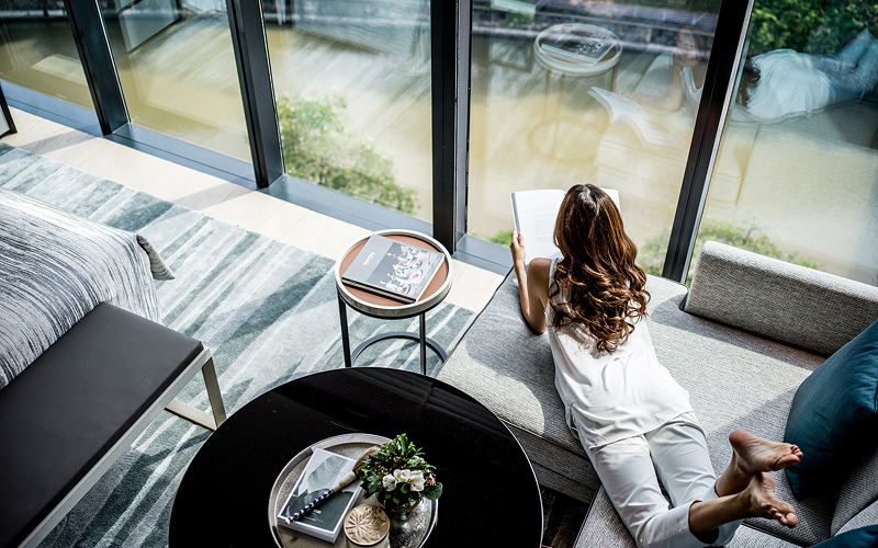 Woman relaxing and reading a book by a floor-to-ceiling window.
