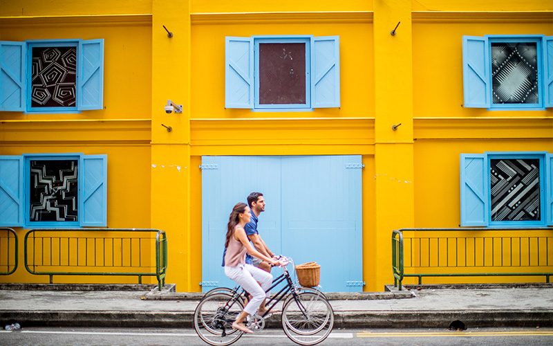 Two people cycling past yellow building.